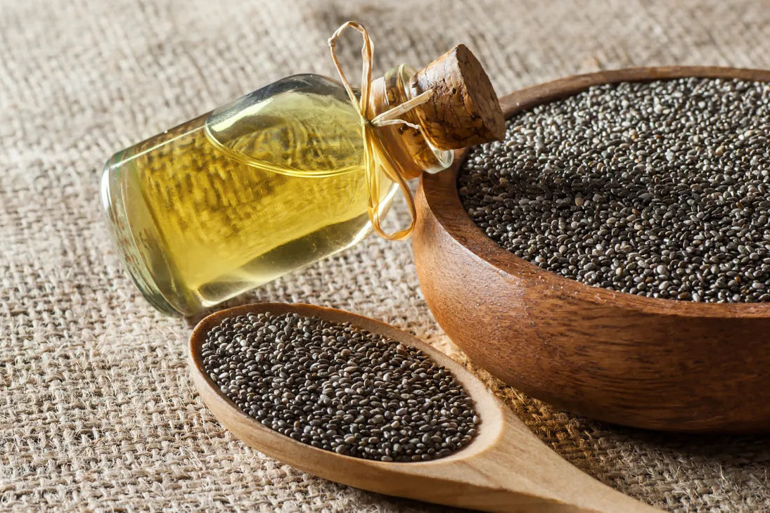 Bottle of chia seed oil with a wooden spoon and bowl filled with chia seeds on a textured fabric background.