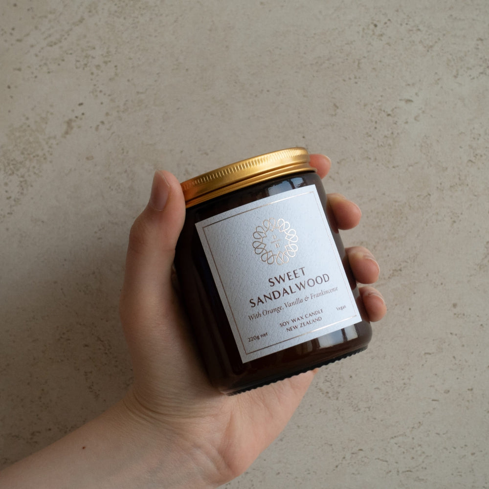 Hand holding a jar of 'Sweet Sandalwood' candle against a neutral background